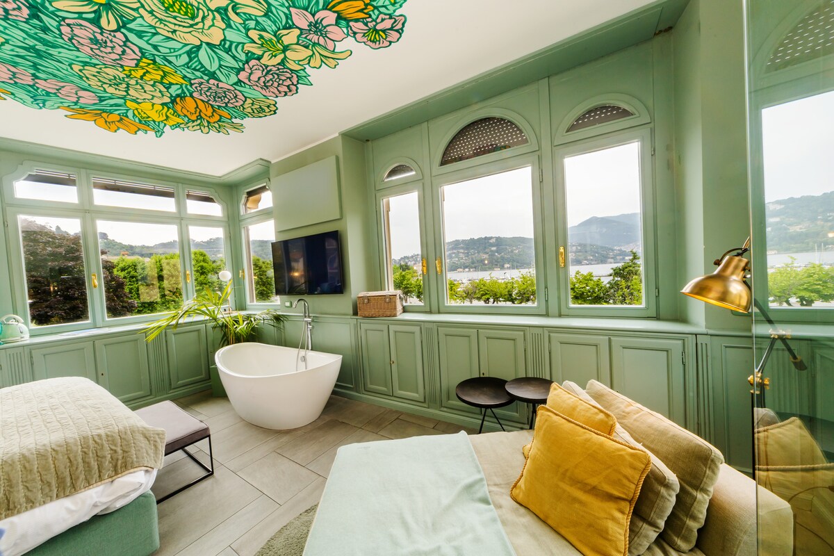 A light-filled room features large windows offering a panoramic view of the lake. A freestanding bathtub with modern fixtures sits near the windows, complemented by a comfortable seating area adorned with colorful cushions. The ceiling showcases a vibrant floral design, enhancing the space's charm.