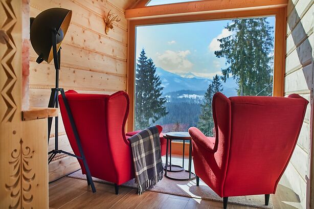 Two red armchairs are positioned near a large window, providing views of the surrounding trees and mountains. A small circular table is situated between the chairs, with a blanket draped over one. Natural light complements the warm wooden interior.