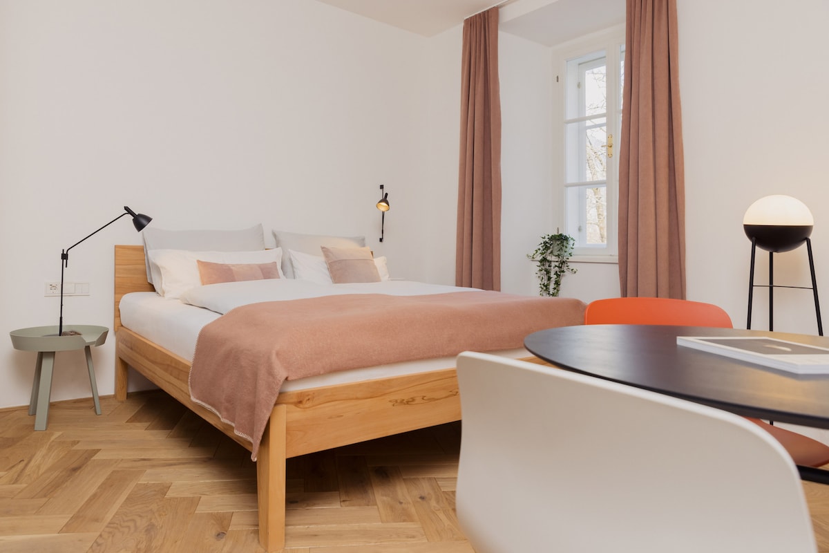 A well-lit bedroom features a spacious bed with light linens, topped with a soft blanket. A small table and chairs provide a cozy dining nook. Natural light filters through the window, enhancing the room's inviting atmosphere.
