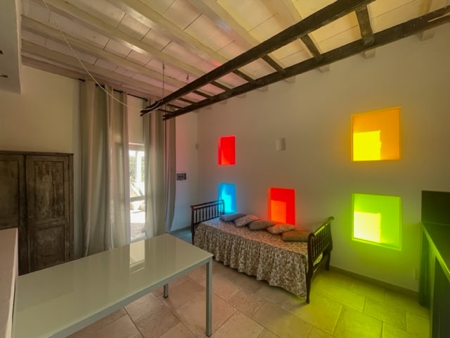 A brightly lit living area features a daybed against a backdrop of colorful window panels in red, orange, and green. A simple table sits nearby, and soft curtains allow natural light to filter into the space. The rustic charm is complemented by exposed wooden beams overhead.