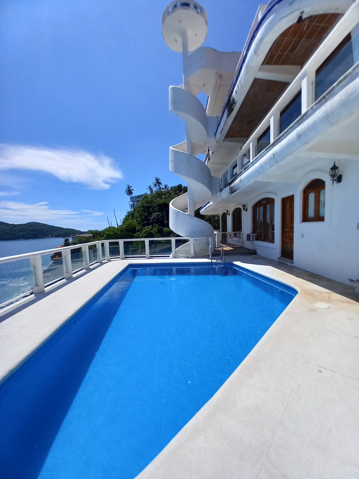 A serene pool area is situated alongside the building, featuring a vibrant blue pool surrounded by light-colored tiles. A spiral staircase is visibly integrated into the architecture, leading up to the upper levels. The tranquil bay and lush greenery create a picturesque backdrop.