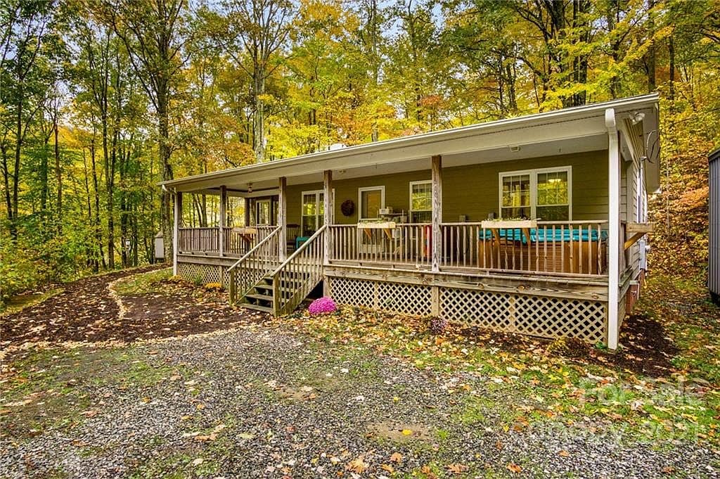 A single-story cabin is displayed amidst a backdrop of vibrant autumn foliage. The porch features a railing and seating area, extending across the front. A pathway leads to the entrance, surrounded by gravel and landscaping, creating a cozy exterior.