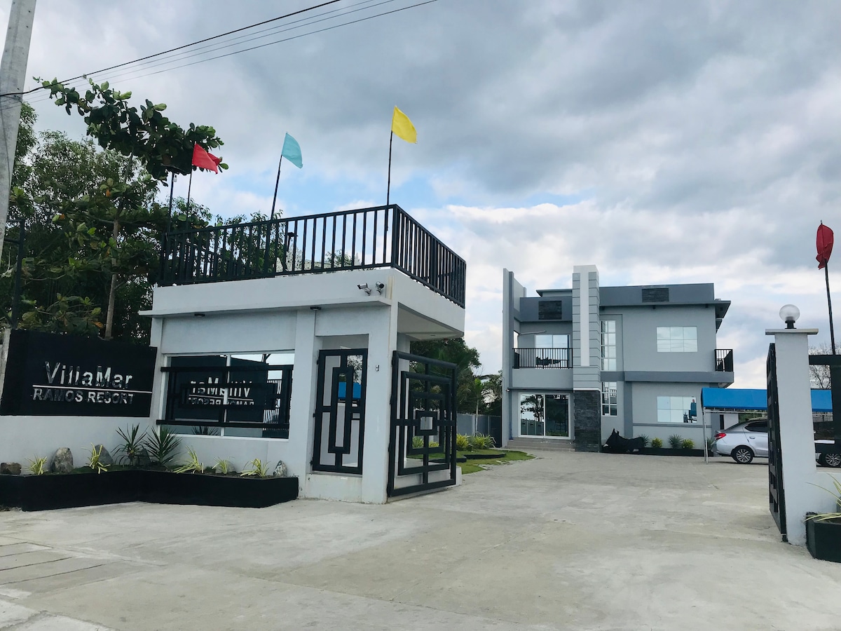 The entrance to VillaMar Ramos Resort is presented, featuring a modern reception area with geometric designs. Flags in varied colors decorate the upper level, while an open driveway connects to the adjacent building. Lush greenery and well-maintained landscaping surround the site.
