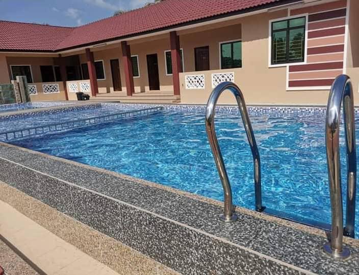 A swimming pool is featured with clear blue water, surrounded by a stone deck. Two stainless steel ladder rails provide access to the pool. The background includes a spacious area with multiple large windows, allowing natural light to enter the space.