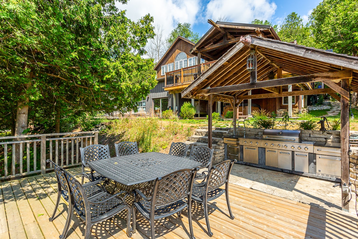 An outdoor dining area features a spacious black table surrounded by eight chairs, situated on a wooden deck. An adjacent outdoor kitchen displays multiple stainless steel grills. Lush greenery and the cottage are visible in the background under a bright blue sky.