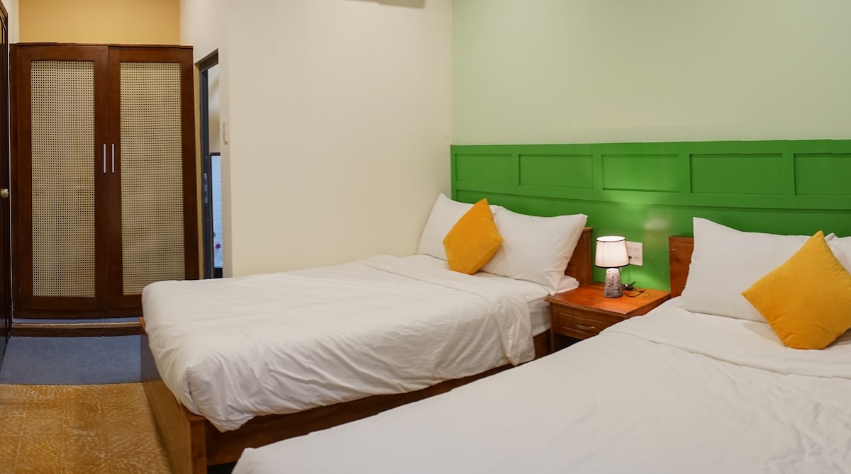 A cozy bedroom is shown with two spacious beds, each adorned with yellow accent pillows. Soft white linens create a fresh appearance. A wooden nightstand and lamp complement the design, while a vibrant green accent wall adds a touch of color. Natural light filters through an adjacent doorway.