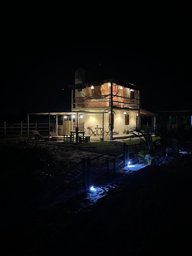 The charming two-story house is illuminated at night, with warm lights glowing from the exterior and patio areas. Surrounding landscaping is softly lit, creating a welcoming ambience. A wooden dining table is visible on the patio, enhancing the outdoor experience.