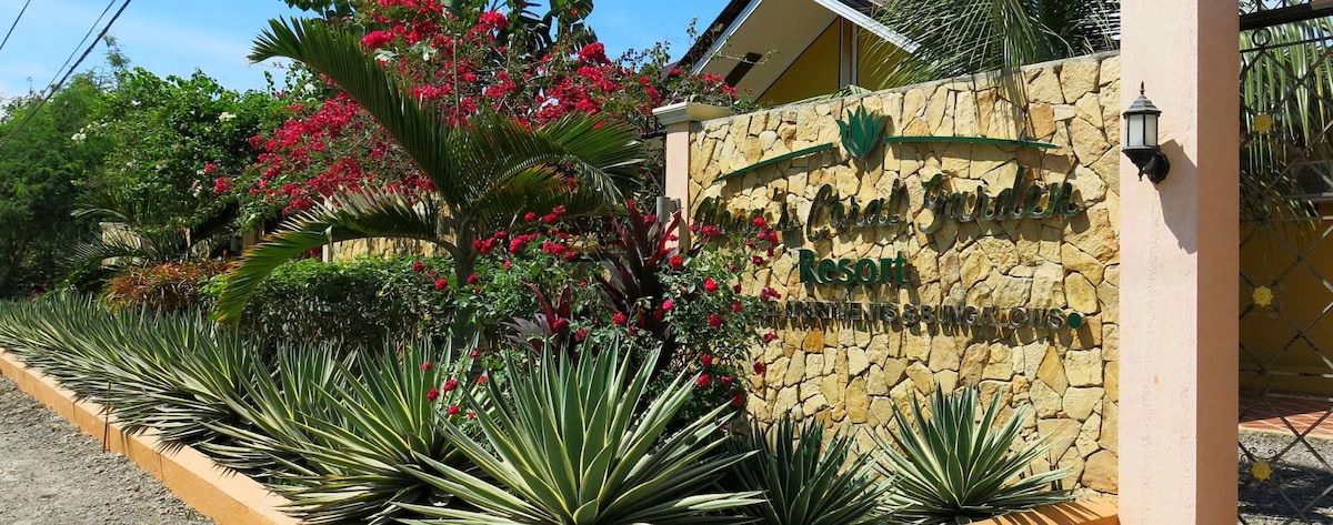 A welcoming entrance to the resort is framed by vibrant greenery and flowers. Various plants, including tall, spiky agave, create a natural border alongside a stone wall displaying the resort's name, emphasizing a tropical feel.