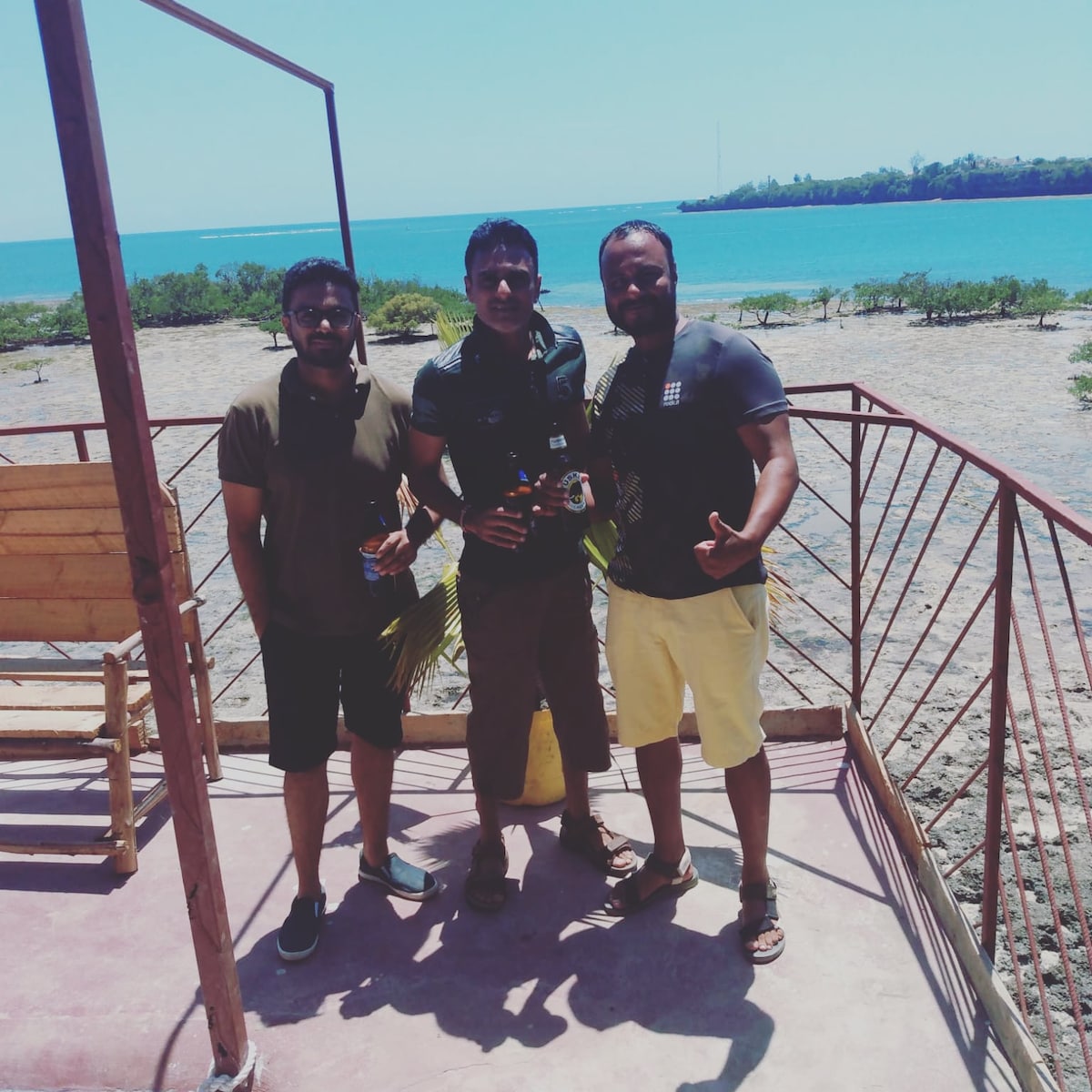 Three individuals stand on a deck overlooking the ocean, with a coastal view in the background. The deck features a simple wooden chair and metal railings. The vibrant blue water is visible, complemented by greenery along the shoreline.
