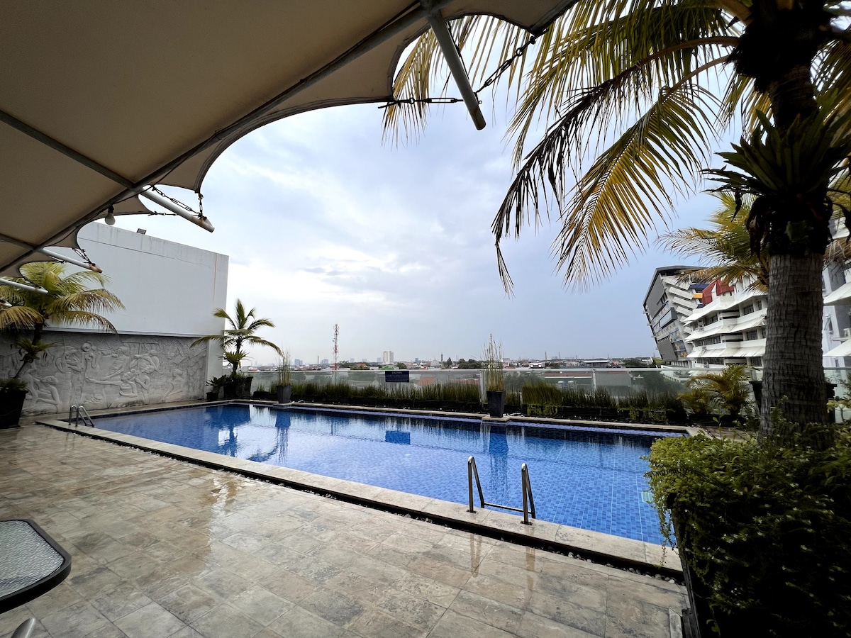 An expansive outdoor area is highlighted, featuring a serene swimming pool bordered by palm trees. The pool's clear water reflects the overcast sky, while modern buildings can be seen in the background, contributing to the urban atmosphere.
