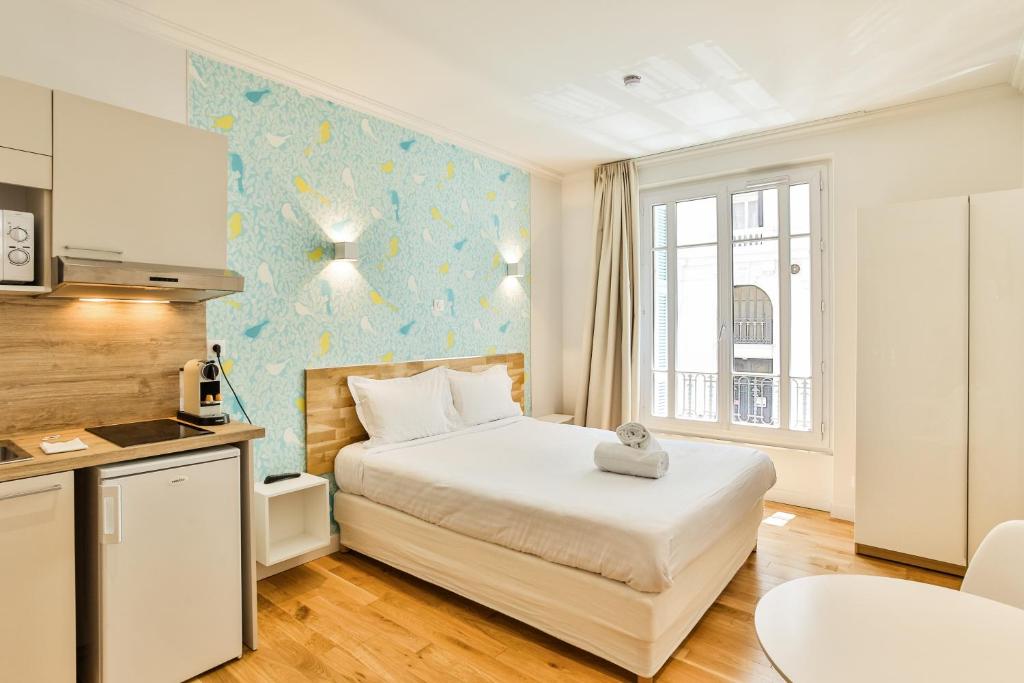 A soothing studio space showcases a spacious bed with fresh linens and a decorative towel placed atop. The kitchen area features modern appliances, and light wooden accents complement the colorful wall. Natural light streams in through the large windows, enhancing the overall ambiance.