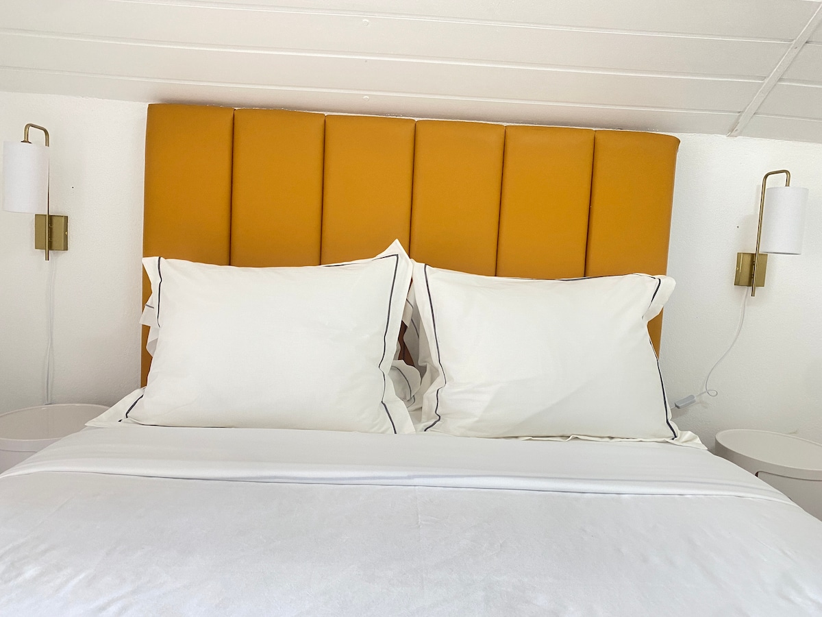 A comfortable sleeping area is highlighted by a distinctive orange headboard. Two neatly arranged pillows rest against the headboard, while soft bedside lamps provide gentle illumination. Minimalist nightstands are positioned on either side of the spacious bed, creating a serene environment.