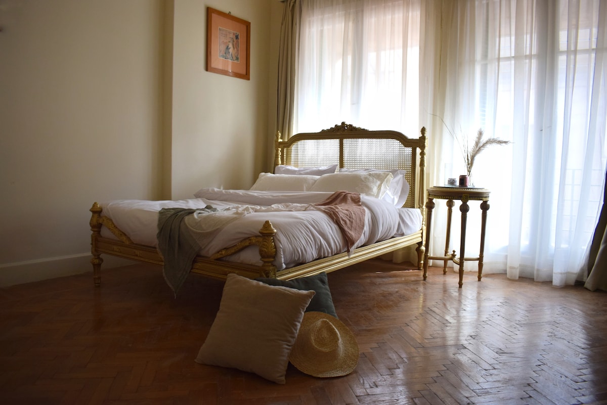 A spacious bedroom is highlighted by a ornate golden bed frame with neatly arranged bedding. Natural light filters through sheer curtains, illuminating a small round table and a decorative pillow on the floor. A straw hat rests casually nearby.