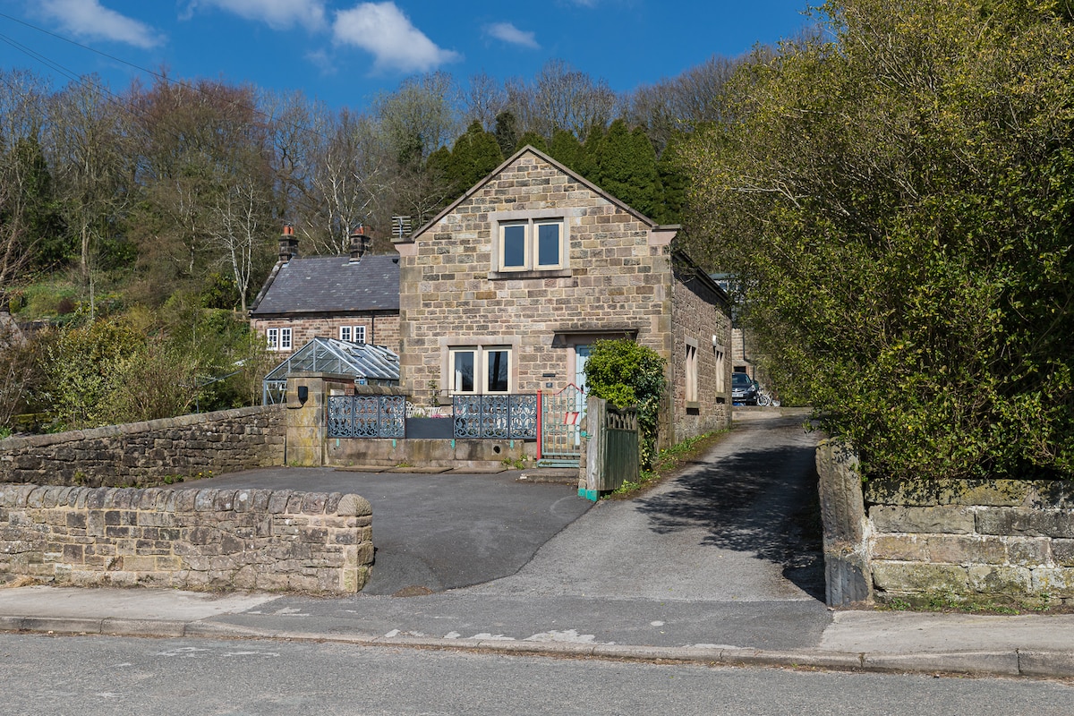 A stone cottage is set against a backdrop of greenery, with a gravel driveway leading to the entrance. Sunlight bathes the facade, highlighting the rustic charm and traditional architecture of the home. Nearby trees add to the serene village atmosphere.
