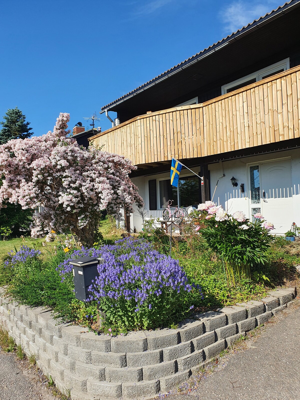 The exterior of a two-story villa features a lush garden with blooming pink and purple flowers, alongside a decorative mailbox. A wooden balcony extends from the upper floor, adorned with a flag. Bright blue skies enhance the welcoming appearance of the home.