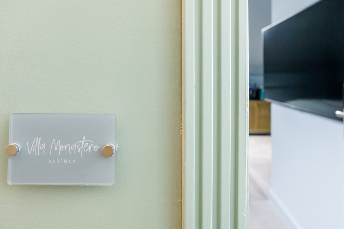 A clear sign indicating 'Villa Monastero Varenna' is displayed on a light green wall. In the background, a dark television unit is partially visible, suggesting a modern living space with minimalistic decor.