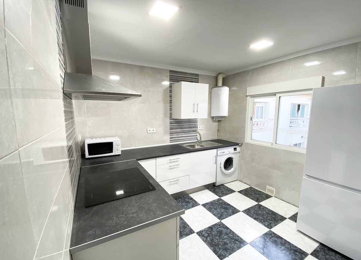 A modern kitchen is displayed, featuring light-colored cabinets and dark countertops. An array of appliances includes a microwave, a refrigerator, and a washing machine. A window allows natural light to enter, illuminating the checkered black and white floor.