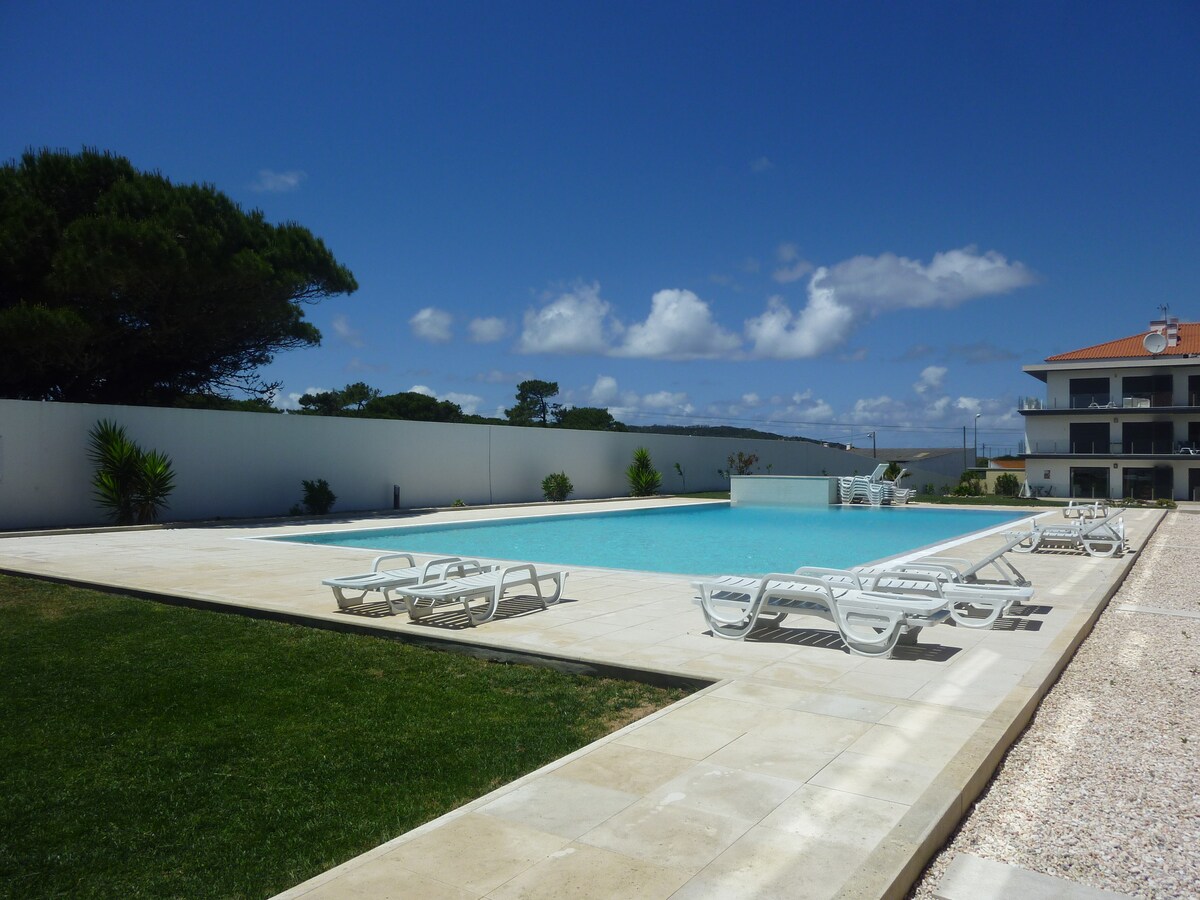 An outdoor swimming pool is surrounded by a paved area, featuring several white lounge chairs for relaxation. Lush green grass borders the pool, and a tall tree provides shade. The building adjacent to the pool displays a modern architectural design under a clear blue sky.