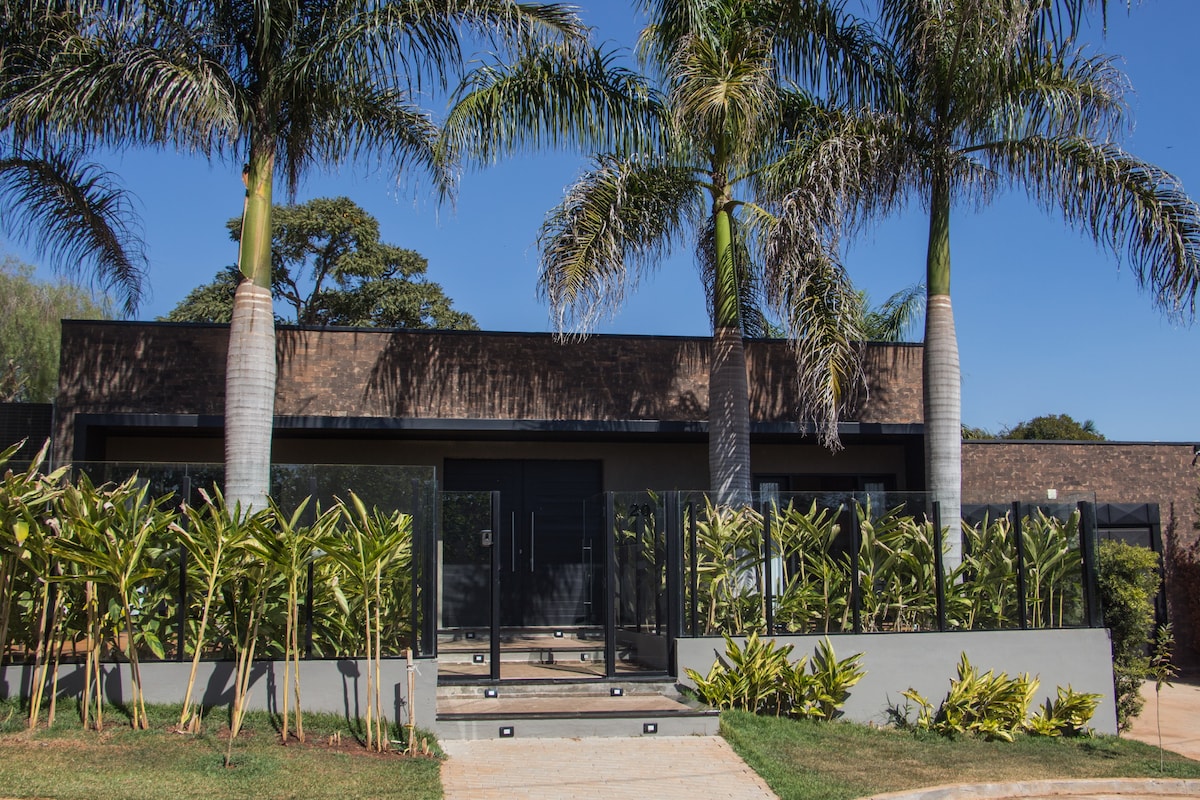 A modern house is framed by tall palm trees, with a clear blue sky above. Lush greenery outlines the landscaped entrance, featuring wide steps leading to glass doors. A sleek, contemporary exterior highlights the architectural design.