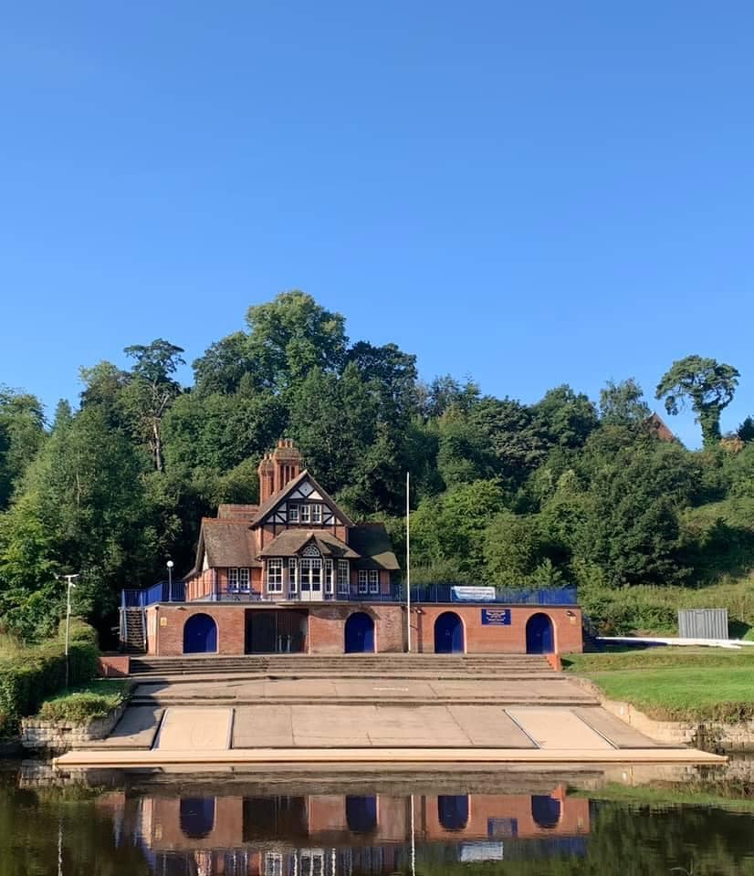 A charming riverside building is showcased against a clear blue sky. The structure features a mix of brick and timber elements, complemented by surrounding trees that add greenery to the landscape. Reflection in the water enhances the serene setting.