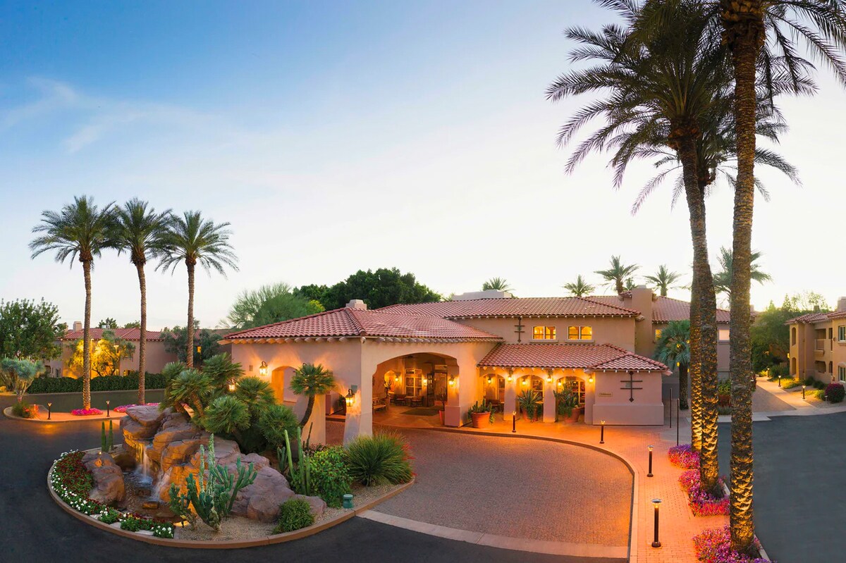 The entrance to the property is framed by well-maintained landscaping, featuring palm trees and colorful flowers. The building, designed with a Southwestern architectural style, has warm lighting that enhances its welcoming ambiance as it blends with the natural surroundings.