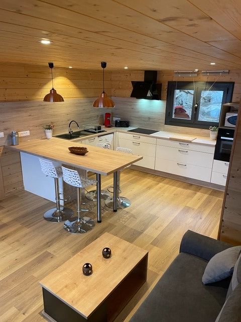 An open kitchen space features modern appliances and ample countertop areas. A wooden dining table with three bar stools provides a casual eating area. Soft lighting from pendant lamps creates a warm ambiance while light wood accents enhance the room's coziness.
