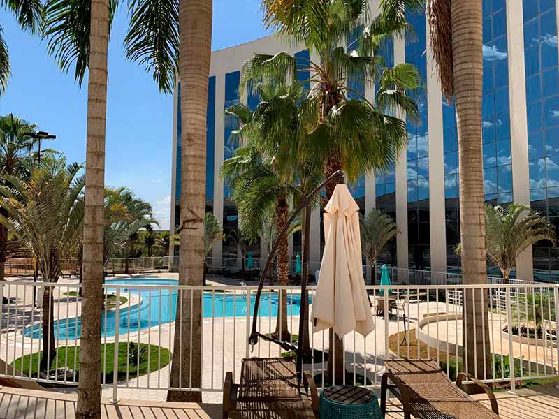 A view of the hotel pool surrounded by palm trees is presented. Lounge chairs, arranged under the shade of large umbrellas, create a relaxing atmosphere. The blue water of the pool reflects the clear sky and the glass facade of the hotel.