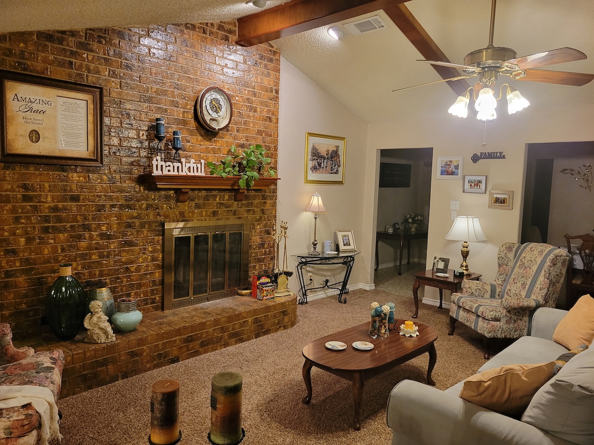 A cozy living room features a brick fireplace with a decorative mantel. Comfortable seating is provided by a floral-patterned chair and a light-colored sofa. A coffee table holds decorative items, and warm lighting is created by lamps placed beside each seating area, enhancing the room's inviting feel.