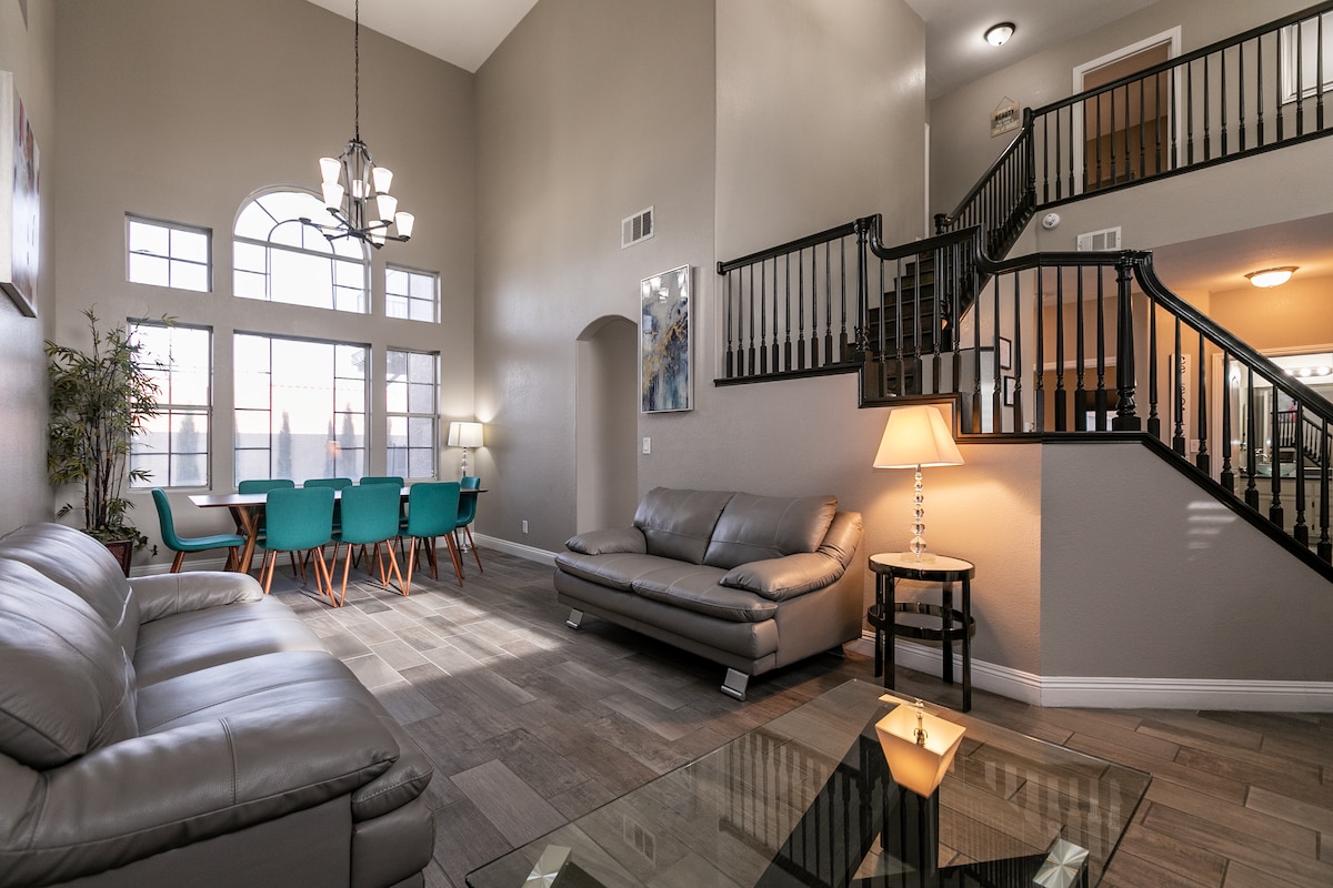An open living area is featured, showcasing sleek leather sofas and a glass coffee table. Large windows allow natural light to fill the space. A formal dining area with teal chairs is visible, while a staircase leads to the upper level, adding depth to the layout.