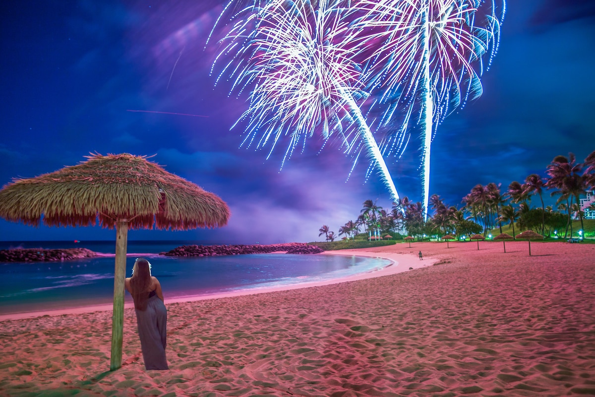 A serene beach scene captures a woman leaning against a thatched umbrella. Colorful fireworks illuminate the night sky above a calm shoreline, with palm trees lining the background and soft sand in the foreground.