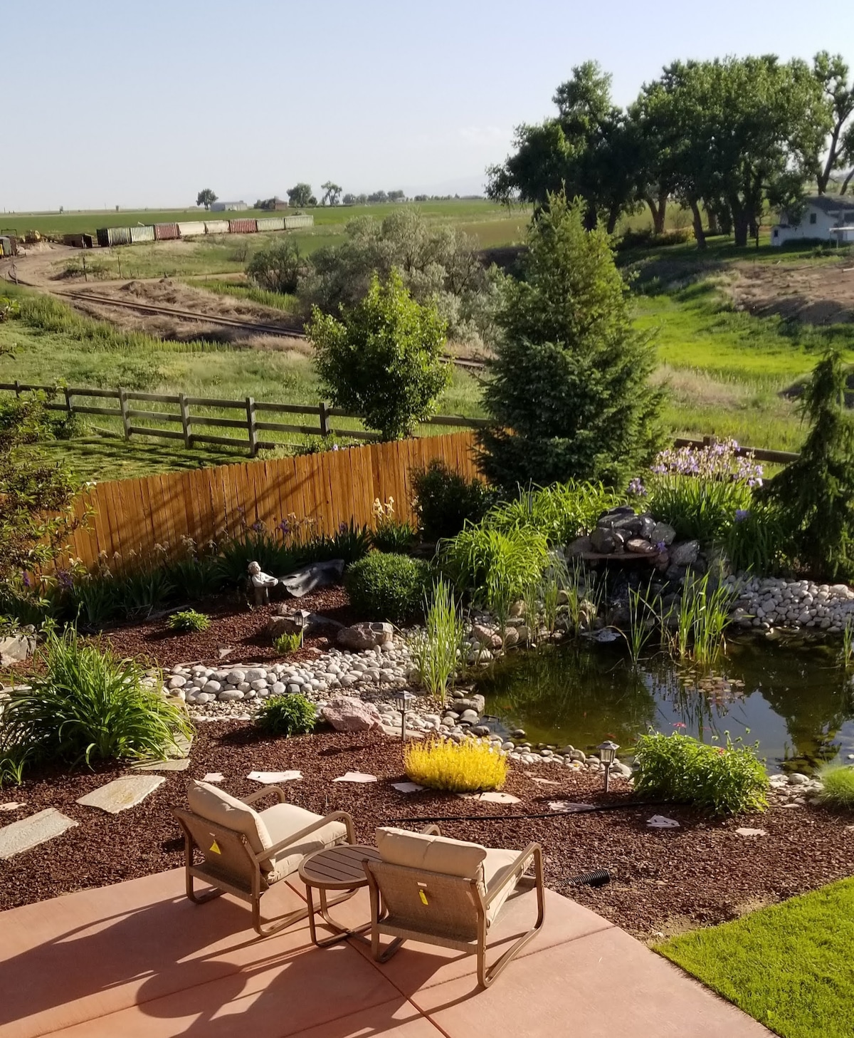 A serene outdoor area features two seating chairs positioned on a patio overlooking a landscaped garden. A pond with rocks and aquatic plants is present, along with a gentle stream and various greenery. A fence lines the area, providing a natural boundary.