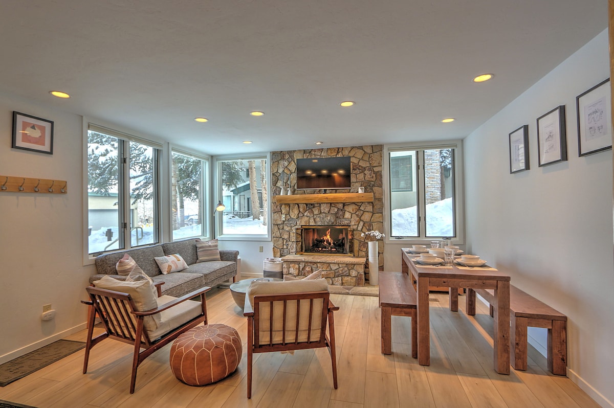 A modern living room features a stone fireplace and large windows allowing natural light. Comfortable seating includes a sofa, wooden chairs, and an inviting area rug. A wooden dining table, suitable for six, is set for meals, enhancing the communal space.