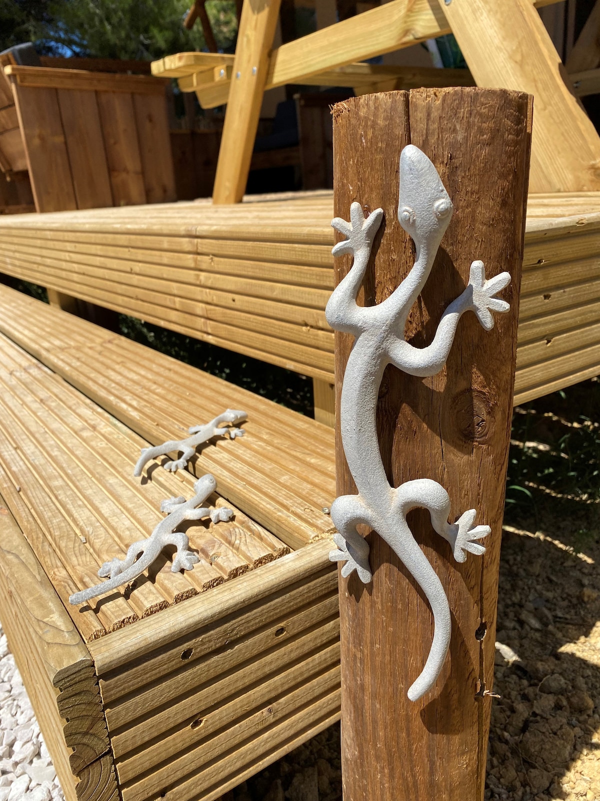 A wooden post is adorned with a stylized lizard sculpture, while additional smaller lizards are positioned nearby on the wooden steps. The natural wooden texture contrasts with the light-colored sculptures, indicating a blend of nature and artistic design.