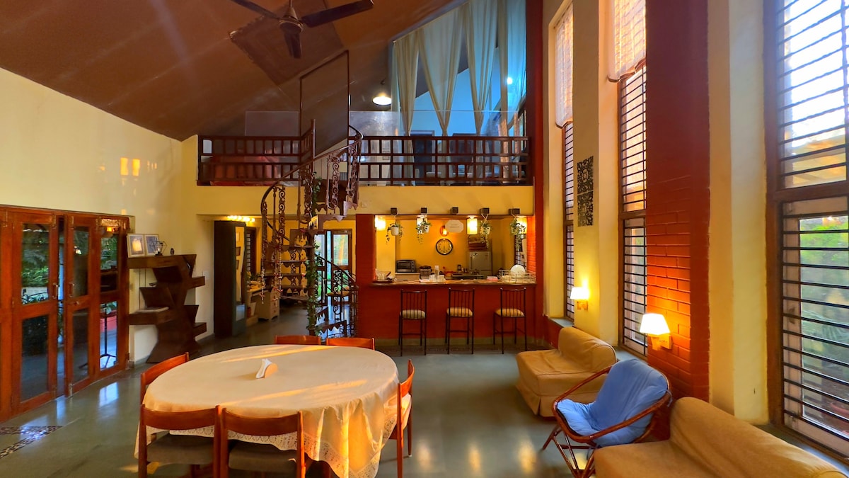 A spacious living room features large windows allowing natural light to fill the space. A circular dining table is placed in the center, surrounded by seating options. An open kitchen is visible in the background, with a staircase leading to the upper level, enhancing the flow of the area.