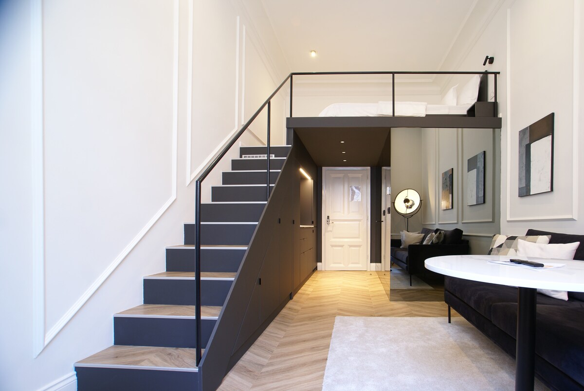 A modern studio apartment features a lofted sleeping area accessible by sleek stairs. The open living space includes a dark sofa and a round table. Light wood flooring adds warmth, while the walls are adorned with framed art and crisp molding, creating an airy atmosphere.