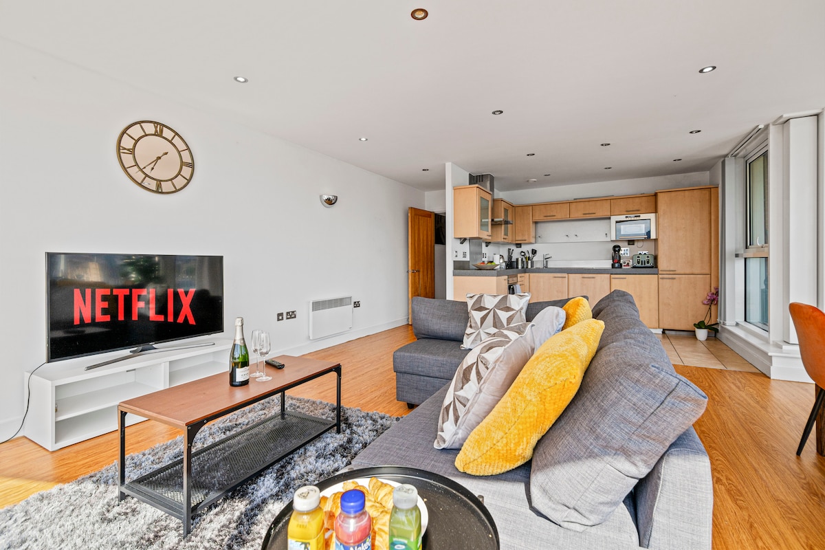 A modern living area features a comfortable seating arrangement with a mix of grey and yellow cushions. A flat-screen TV displays the 'NETFLIX' logo. A wooden coffee table is positioned in front, complemented by a selection of drinks. The kitchen is visible in the background.