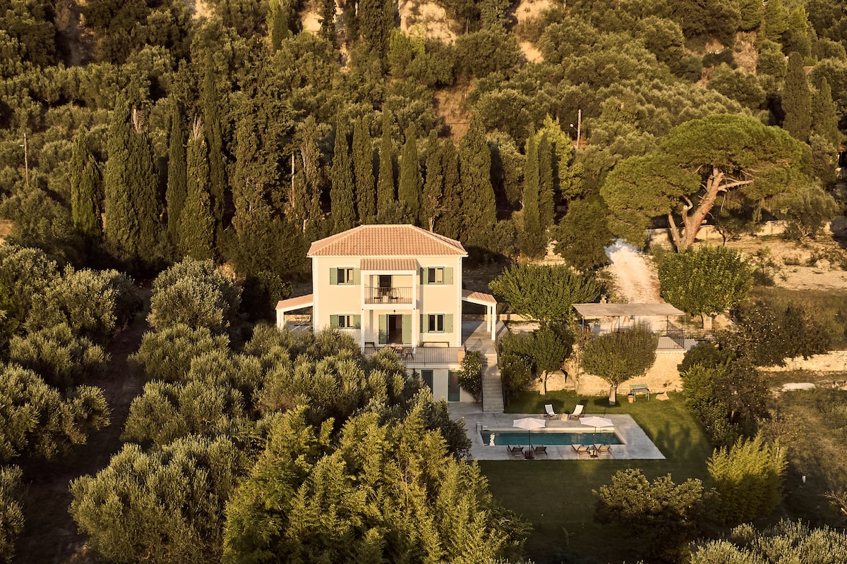 The two-story house is surrounded by lush greenery, featuring cypress and olive trees. A swimming pool is situated in the foreground, complementing the spacious outdoor area with cozy lounging options. The balcony on the second floor offers views of the scenic landscape.