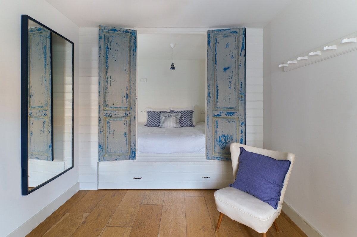 A cozy sleeping area is defined by a built-in bed frame with vintage blue shutters, highlighting a minimalist design. A large mirror reflects natural light, while a small white chair with a purple cushion adds a touch of color to the space.
