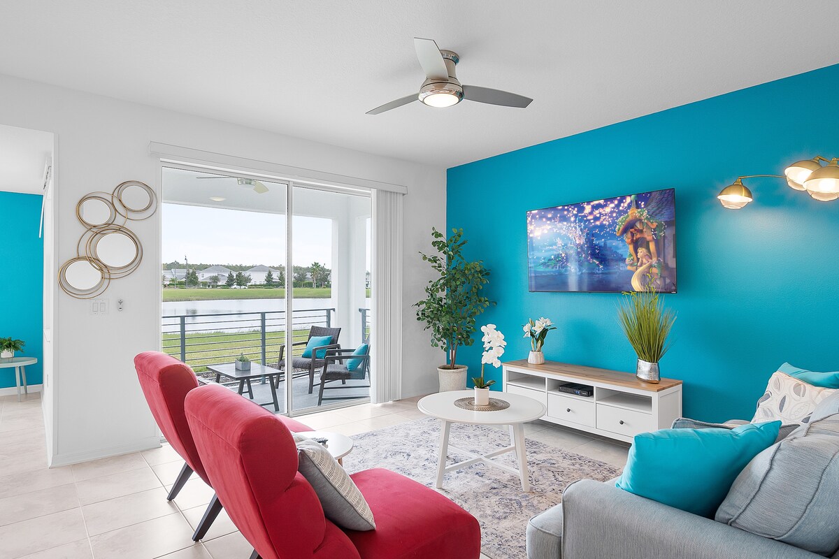 The living area is highlighted by a vibrant blue accent wall, complemented by modern furnishings. A comfortable red chair and light blue couch sit beside a round coffee table, while a wall-mounted TV displays vibrant artwork. Natural light fills the space through sliding glass doors leading to an outdoor patio.