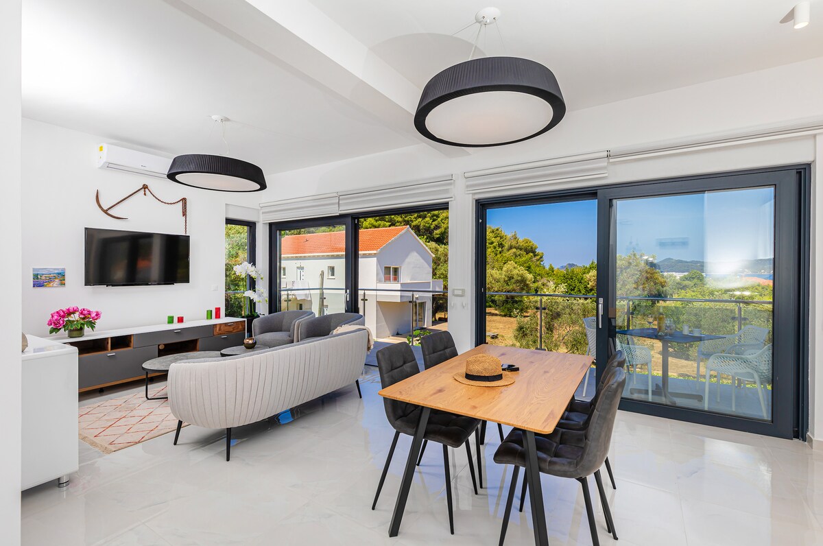 A bright living area features large glass doors that provide views of the surrounding greenery. Comfortable seating is arranged with a modern dining table and six chairs positioned near a wall-mounted television. The space is complemented by stylish light fixtures and light-colored flooring.