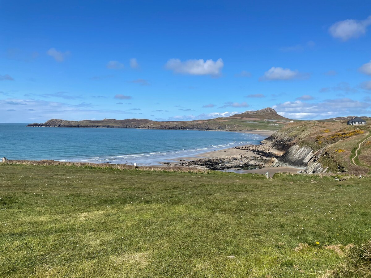 A serene landscape showcases a wide sandy beach bordered by gentle waves. Rolling green hills rise in the distance, under a clear blue sky with scattered clouds. The coastline is viewed from a grassy area, indicating proximity to the shore and natural beauty.