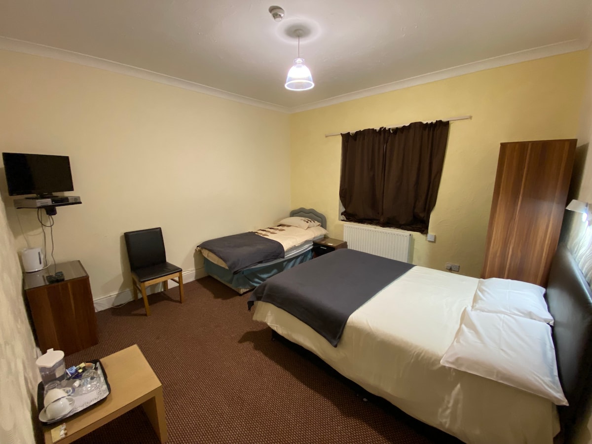 A well-furnished double room features a spacious bed alongside a single bed. A television is mounted on the wall, and a small chair and table are present. Light pours in through a window, partially covered by a dark curtain, enhancing the room's simplicity.