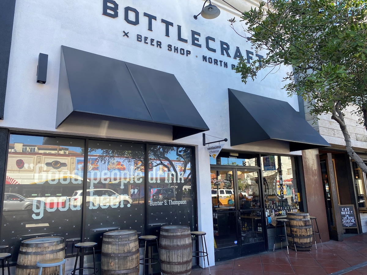 The exterior of a brewery features a modern facade with large glass windows displaying seating areas. Wooden barrels serve as tables alongside the entrance, which is shaded by black awnings. A welcoming sign quotes Hunter S. Thompson, contributing to the inviting atmosphere.