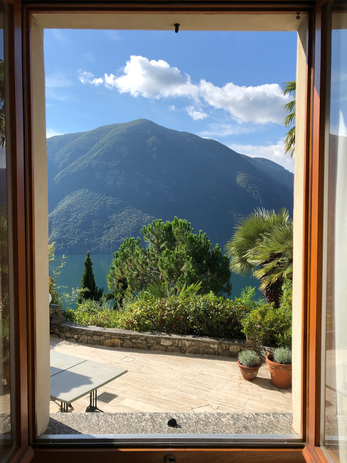 A large window frames a stunning view of lush green mountains and tranquil lake Lugano. The window opens to a stone terrace adorned with potted plants, allowing natural light to illuminate the interior. The scene captures a serene and scenic landscape.