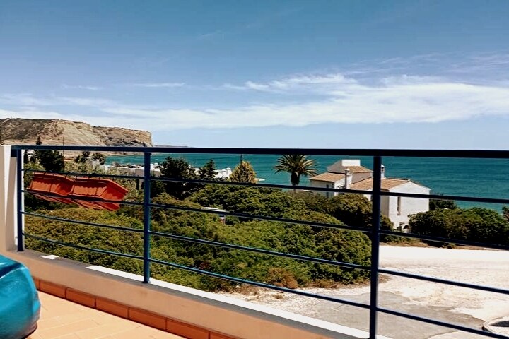 A panoramic view of the coastline is presented from a balcony, featuring the ocean and nearby cliffs. Lush green vegetation is visible in the foreground, along with white buildings nestled along the shore. The sky is clear with a few scattered clouds.