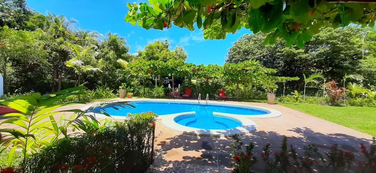 A private pool is framed by vibrant greenery and colorful flowers, creating a serene outdoor oasis. Lounging chairs are positioned around the pool, inviting relaxation under a clear blue sky. The surrounding landscape features lush trees and landscaped areas, enhancing the tranquil environment.