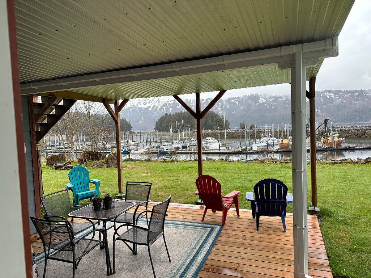 An outdoor patio area is furnished with colorful Adirondack chairs and a round dining table, providing a space for relaxation. The view reveals a boat harbor with several boats docked, framed by green grass and the backdrop of mountains.