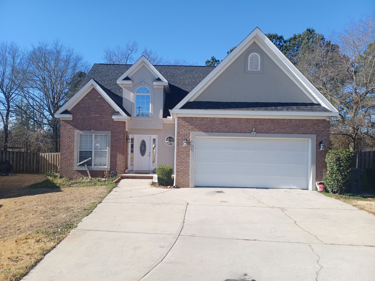 A two-story house with brick and siding exterior is featured, showcasing a driveway leading to a two-car garage. The front yard is open with grass and a fence providing privacy. Large windows flank the entrance, enhancing the inviting facade.