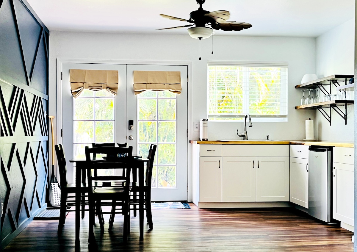 A light-filled kitchen area features a dining table set for four with chairs. Bright cabinetry and a wooden countertop are complemented by a small sink and appliance area, including a refrigerator. Two sets of glass doors open to a lush outdoor view.