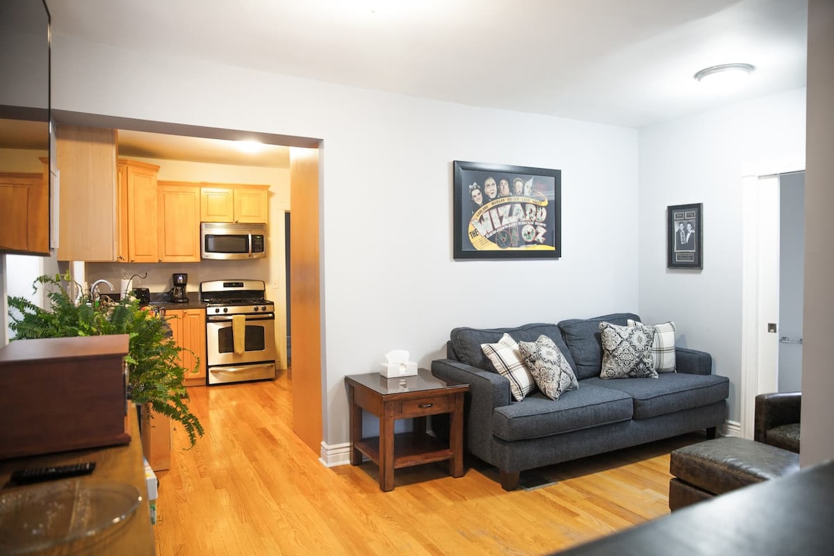 A cozy living area features a comfortable blue sofa adorned with decorative pillows, positioned beside a small wooden side table. The kitchen, visible in the background, showcases warm wood cabinetry and stainless steel appliances, contributing to a welcoming atmosphere.